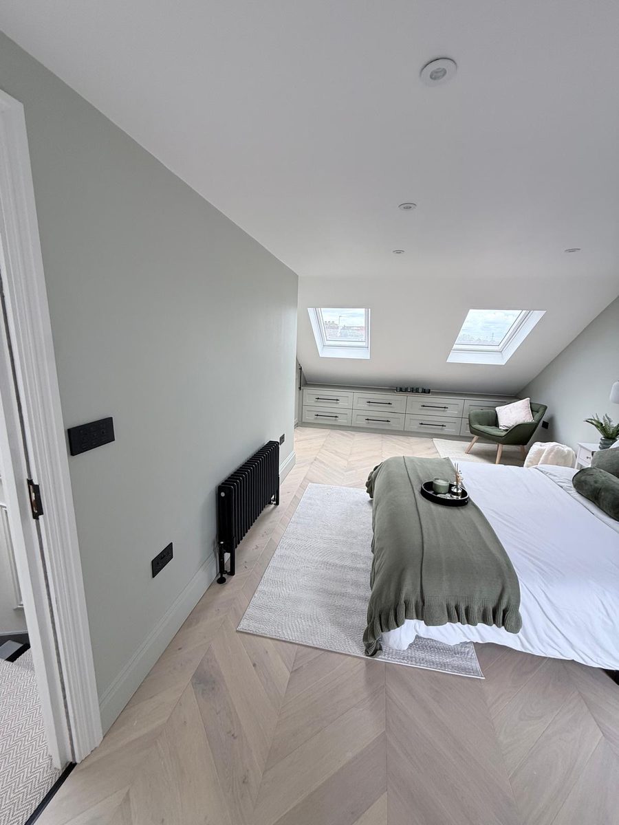 A finished loft bedroom in East London with sage-green walls, oak chevron parquet, fitted wardrobes and a black column radiator, by Platinum Handy Services.