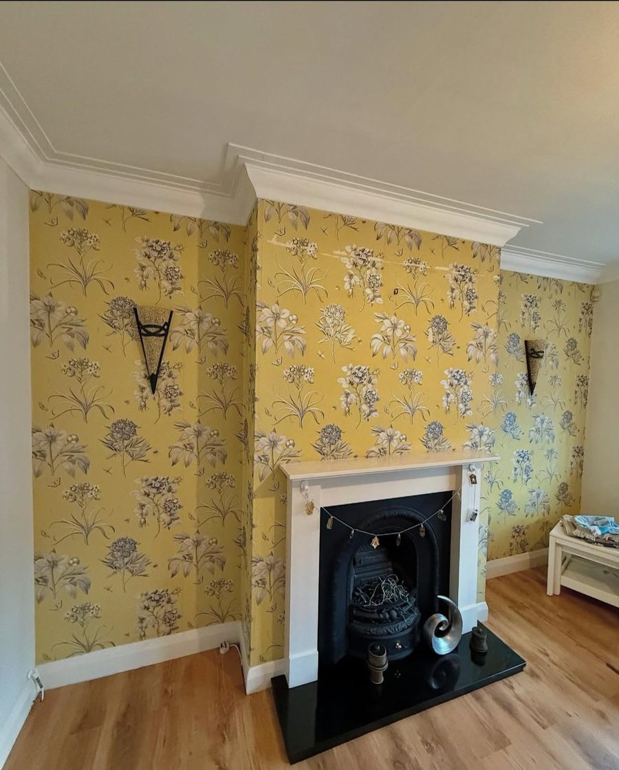 A chimney breast freshly hung with yellow floral feature wallpaper around a period cast-iron fireplace, by Platinum Handy Services.